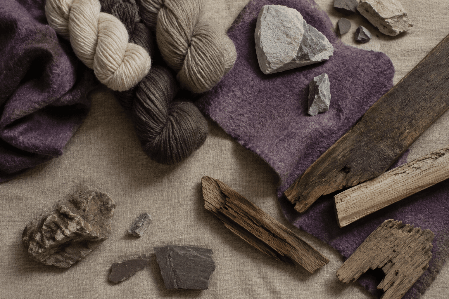 Overhead flat lay of wool yarn, stone fragments, weathered wood and heather felt on a linen workbench